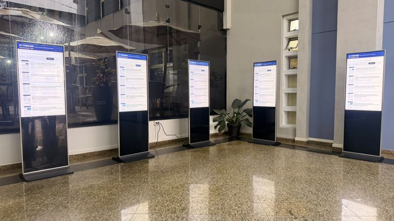 Cinco totens touch de 43 polegadas com a busca de e-pôsteres do Congresso Acadêmico CAAJ aberta na tela, enfileirados no saguão do congresso